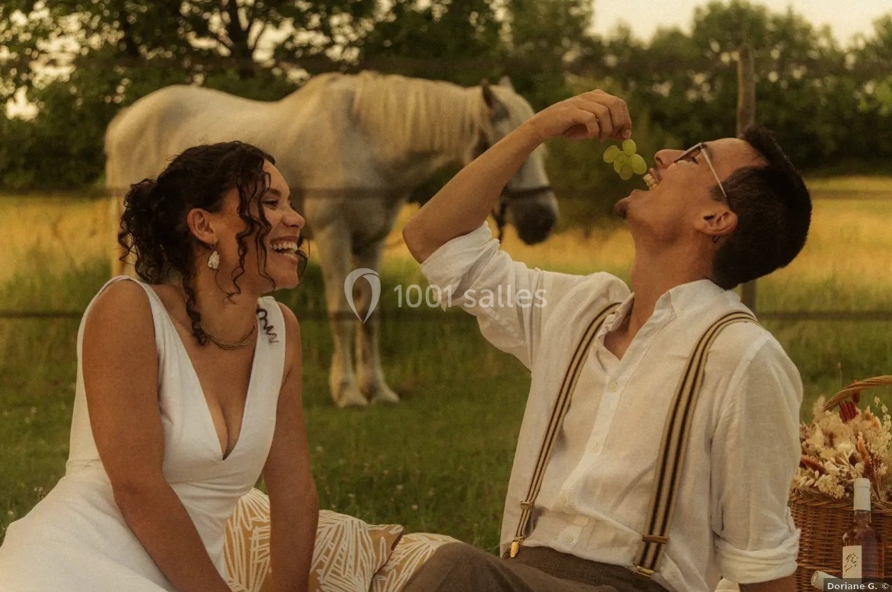 Location salle Saint-Genès-de-Lombaud (Gironde) - Domaine du Château Icla #36 Un couple assis dans un champ partage un moment joyeux, un homme mangeant du raisin, un cheval en arrière-plan.