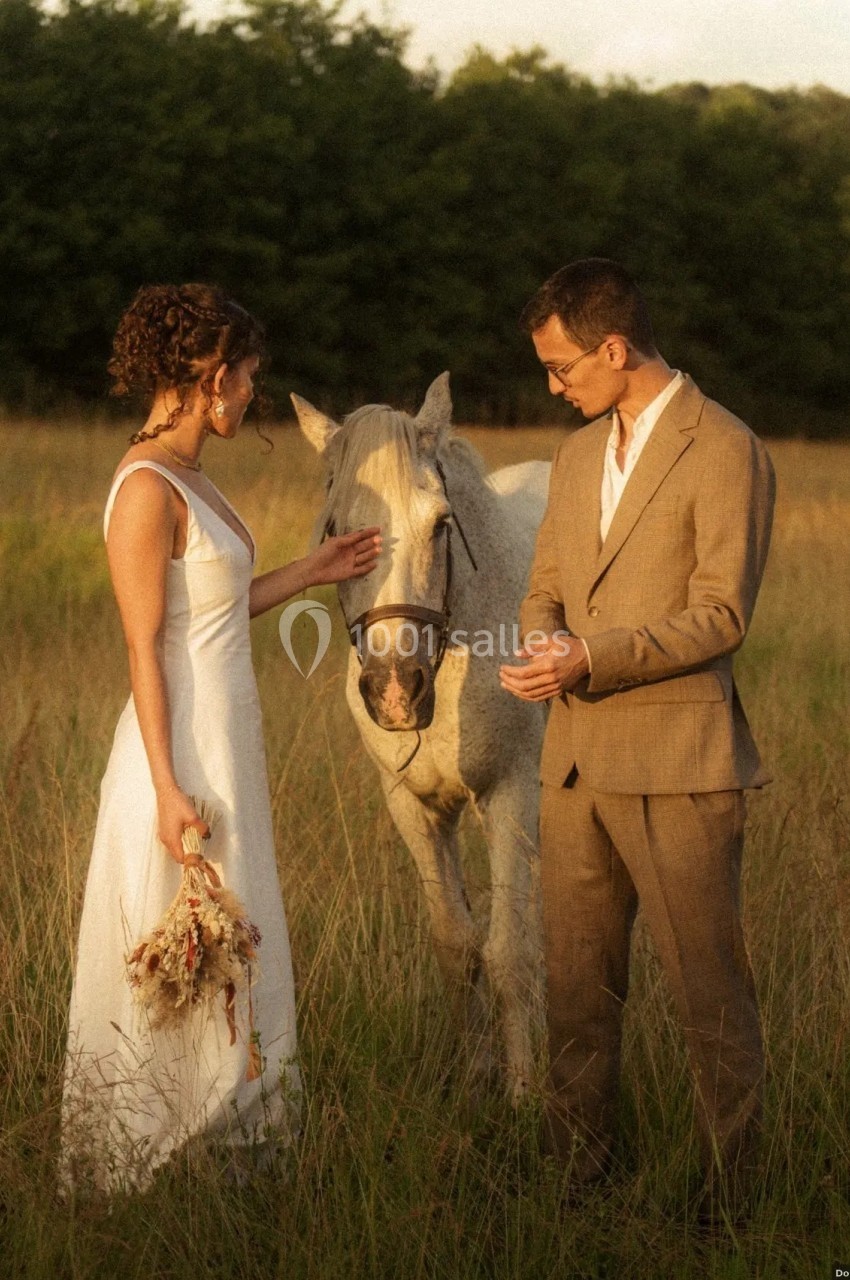 Location salle Saint-Genès-de-Lombaud (Gironde) - Domaine du Château Icla #37 Une femme en robe blanche et un homme en costume beige se tiennent près d'un cheval dans un champ herbeux.