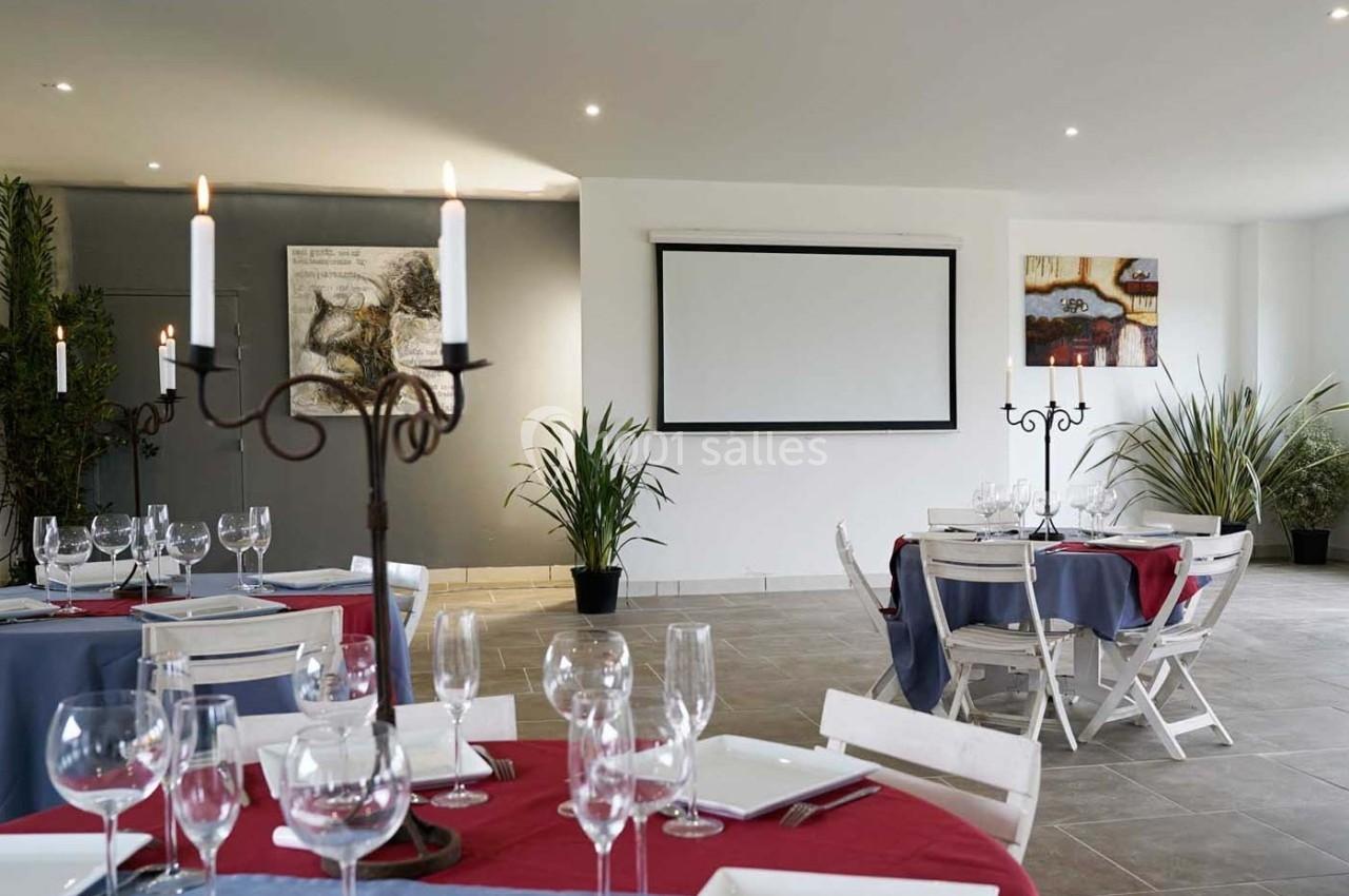 Salle de réception avec tables dressées, nappes colorées, chandeliers, plantes décoratives et écran mural.