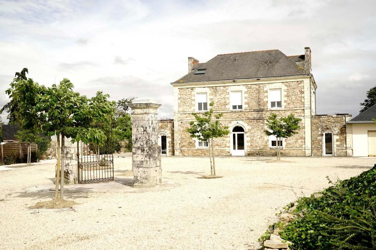 Maison en pierre avec toit en ardoise, entourée d'une cour gravillonnée et de quelques arbres.