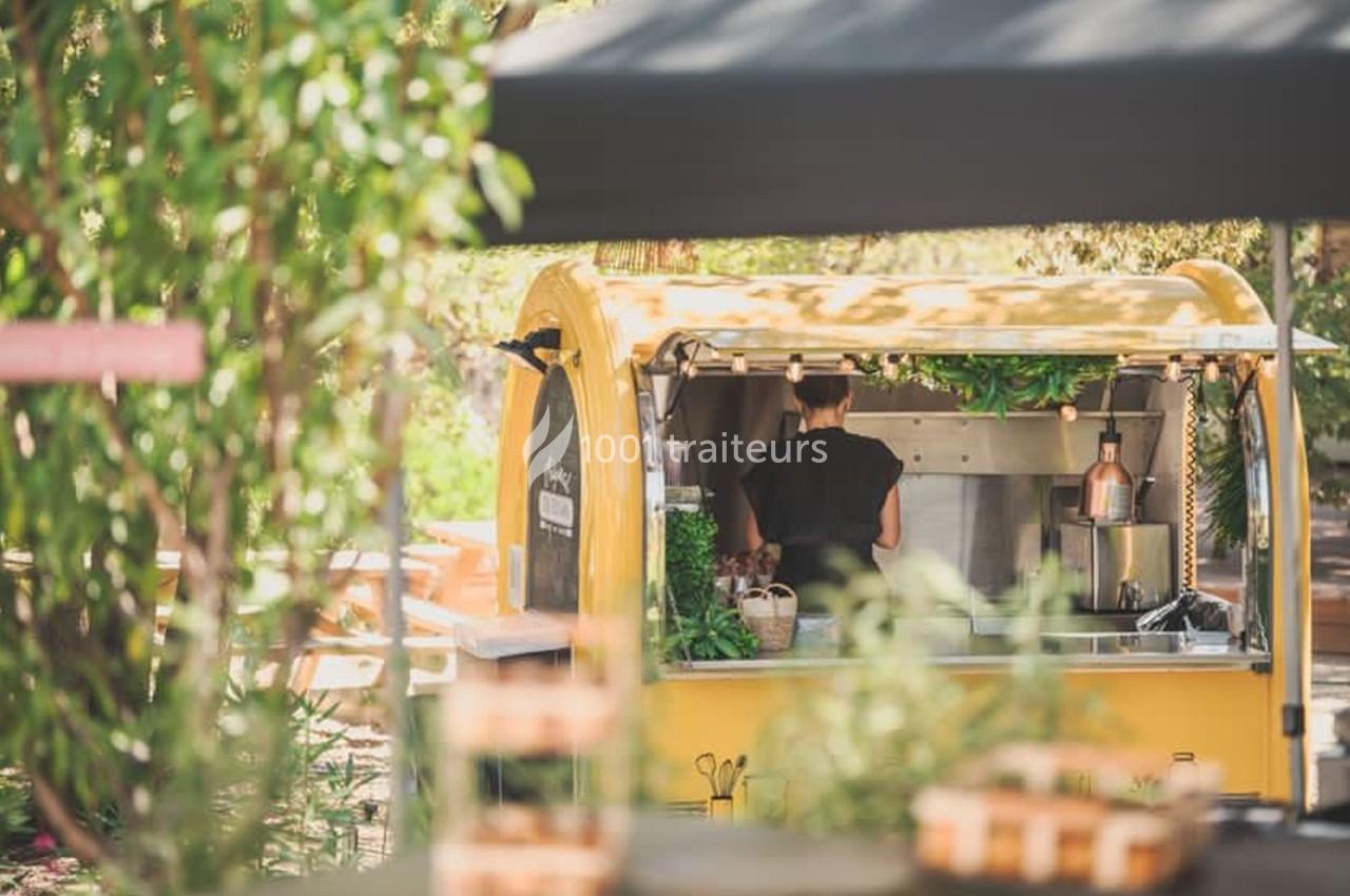 Un food truck jaune dans un cadre extérieur verdoyant, avec une personne préparant des plats à l'intérieur.