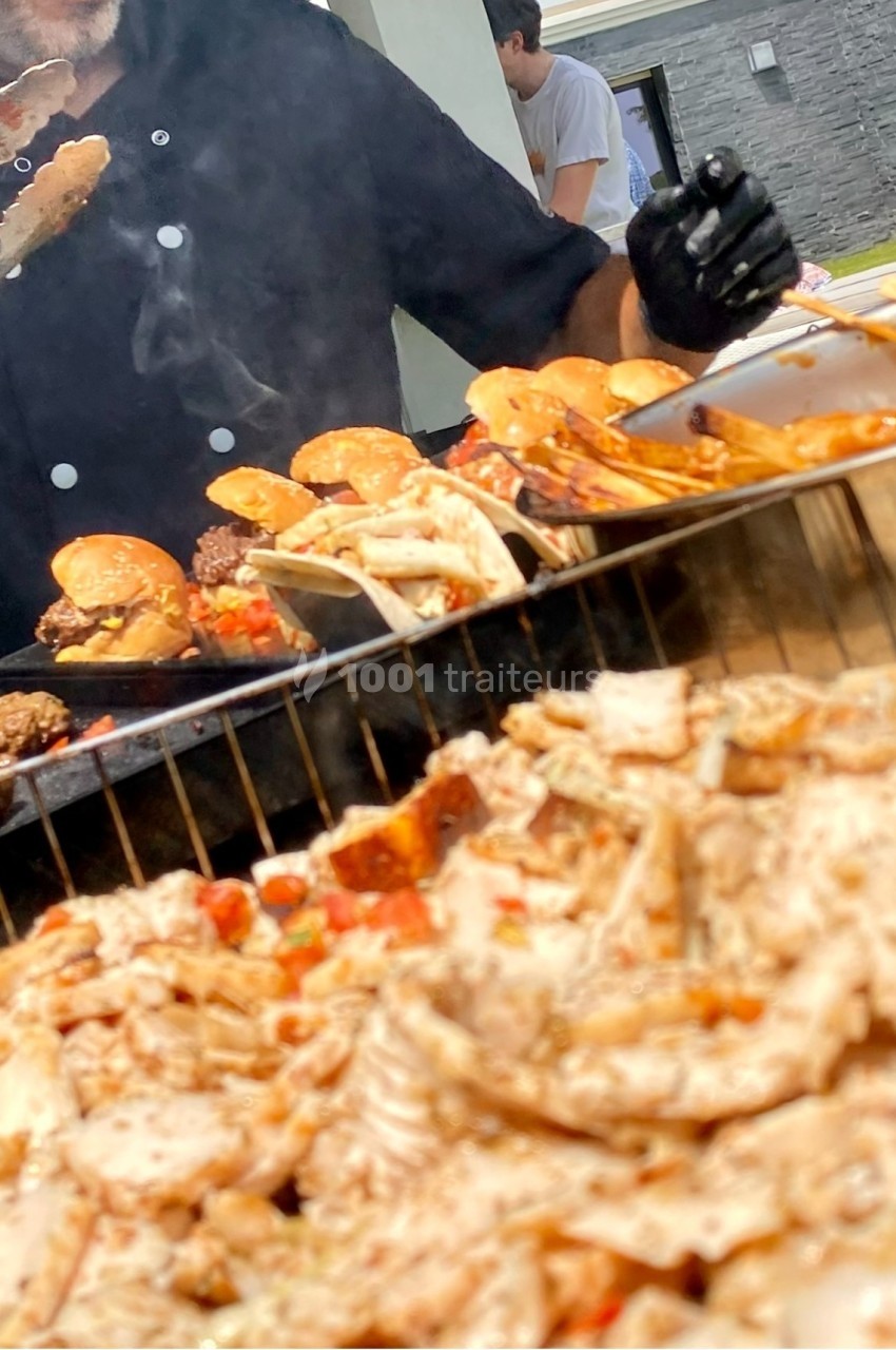 Chef cuisinant des viandes grillées et des burgers sur un barbecue en plein air, avec des convives en arrière-plan.