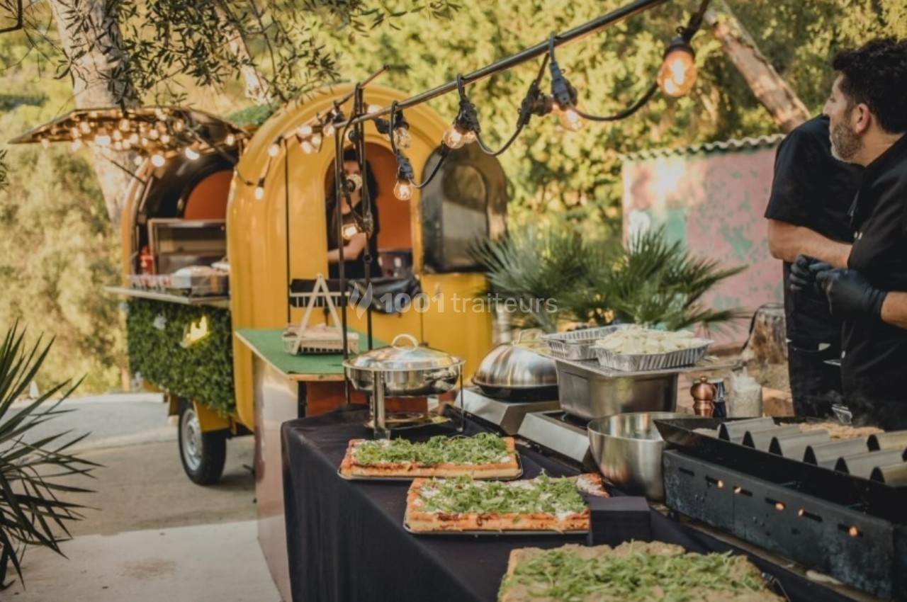 Food truck jaune avec guirlande lumineuse, table garnie de plats variés et cuisinier en arrière-plan, en extérieur.