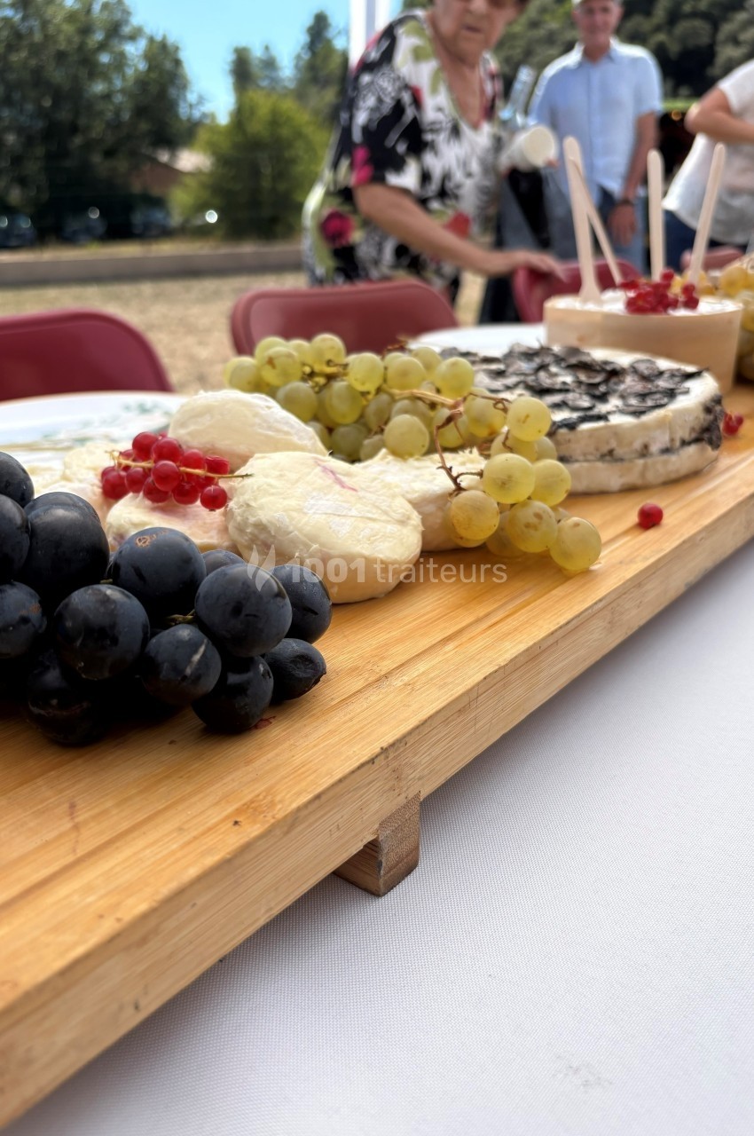 Plateau en bois garni de fromages variés, raisins noirs et blancs, et groseilles, disposé sur une table en extérieur.