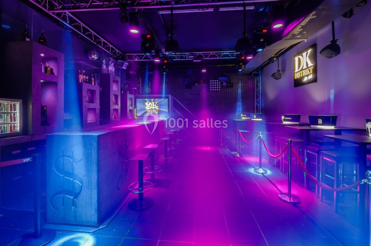Salle de bar moderne avec éclairage coloré, tables hautes, cordons de séparation et ambiance lumineuse violette.