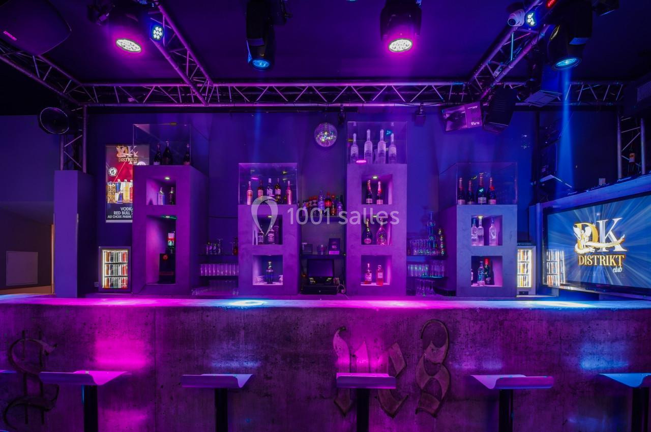 Bar moderne avec éclairage violet et bleu, étagères de bouteilles illuminées et comptoir en béton au premier plan.