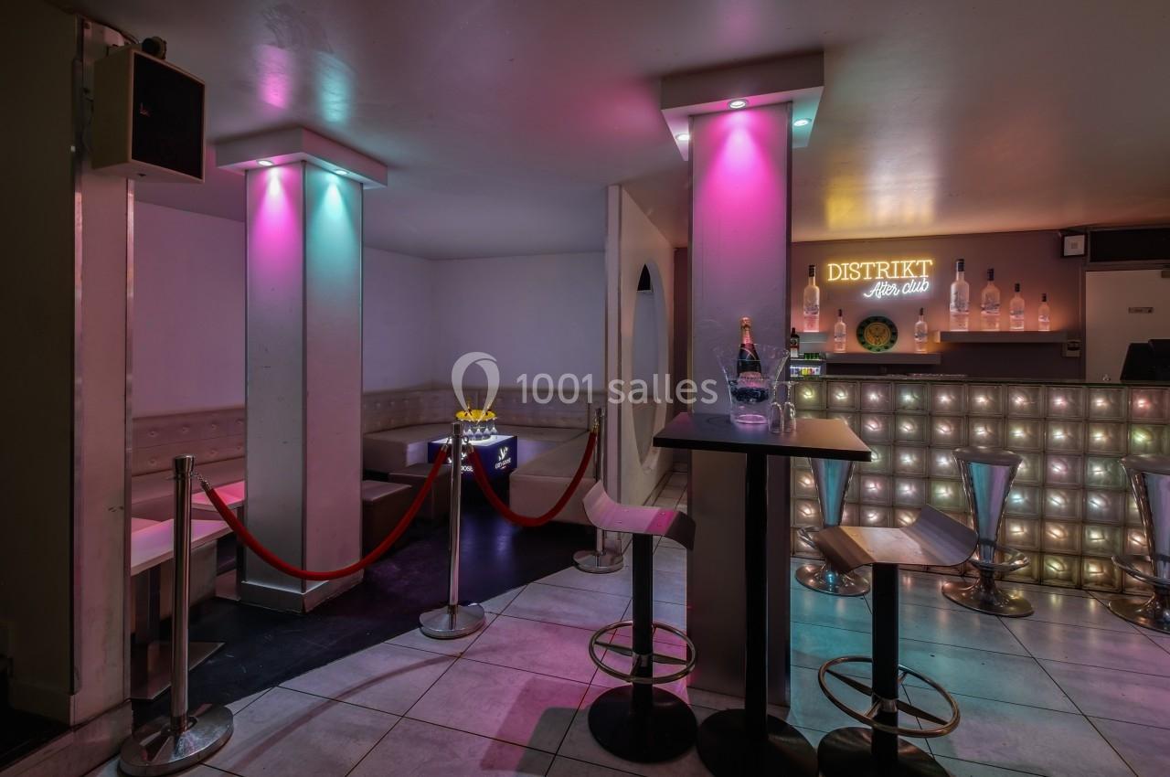 Intérieur d'un bar avec comptoir éclairé, tabourets hauts, table avec bouteille et espace VIP délimité par des cordons…