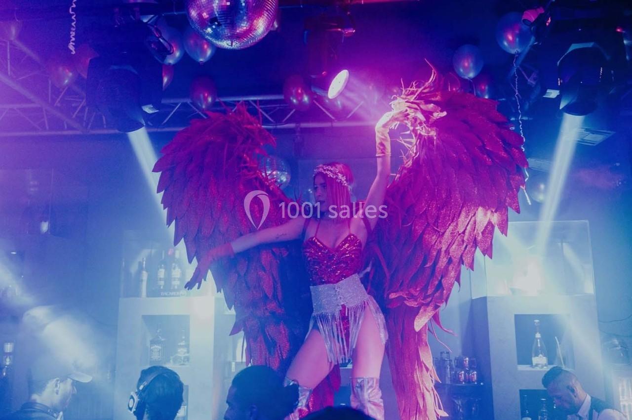 Danseuse en costume rouge à ailes scintillantes sur scène dans une ambiance de boîte de nuit avec éclairage coloré.