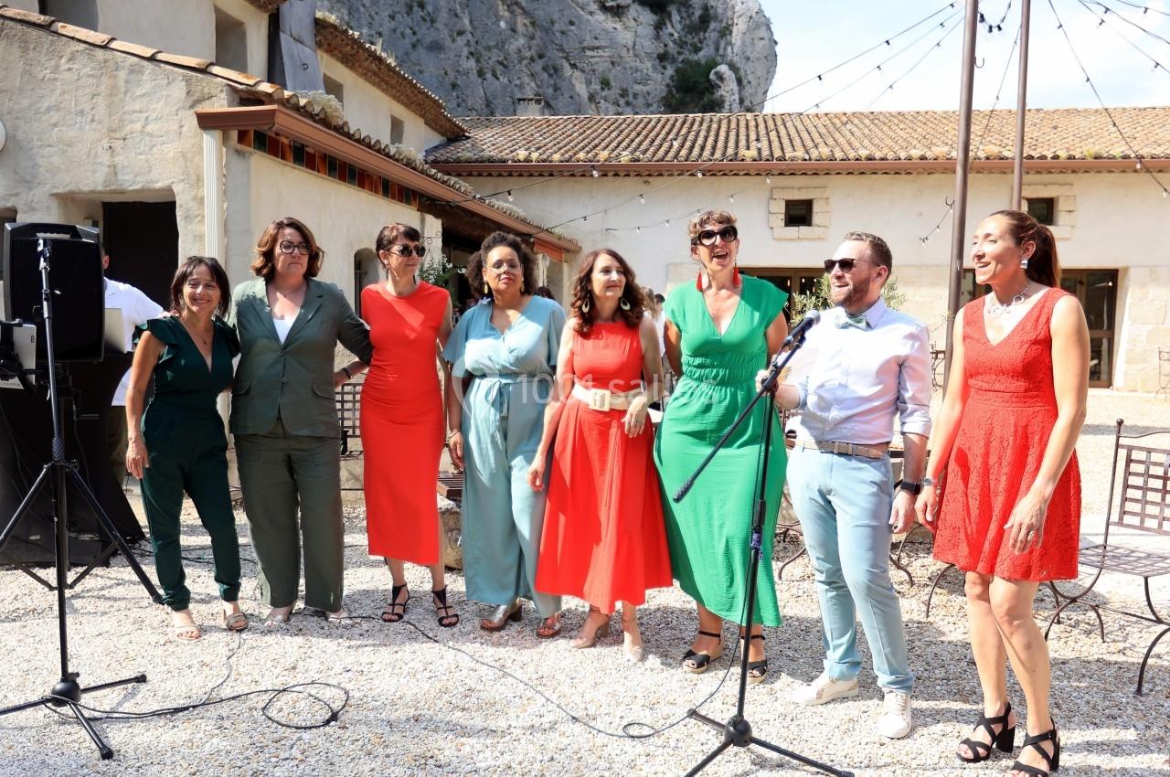 Un groupe de personnes debout en extérieur, souriant devant un micro, dans une cour avec des bâtiments en arrière-plan.