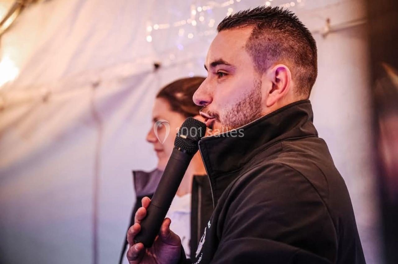 Un homme tenant un micro parle dans un espace éclairé, avec une femme en arrière-plan.