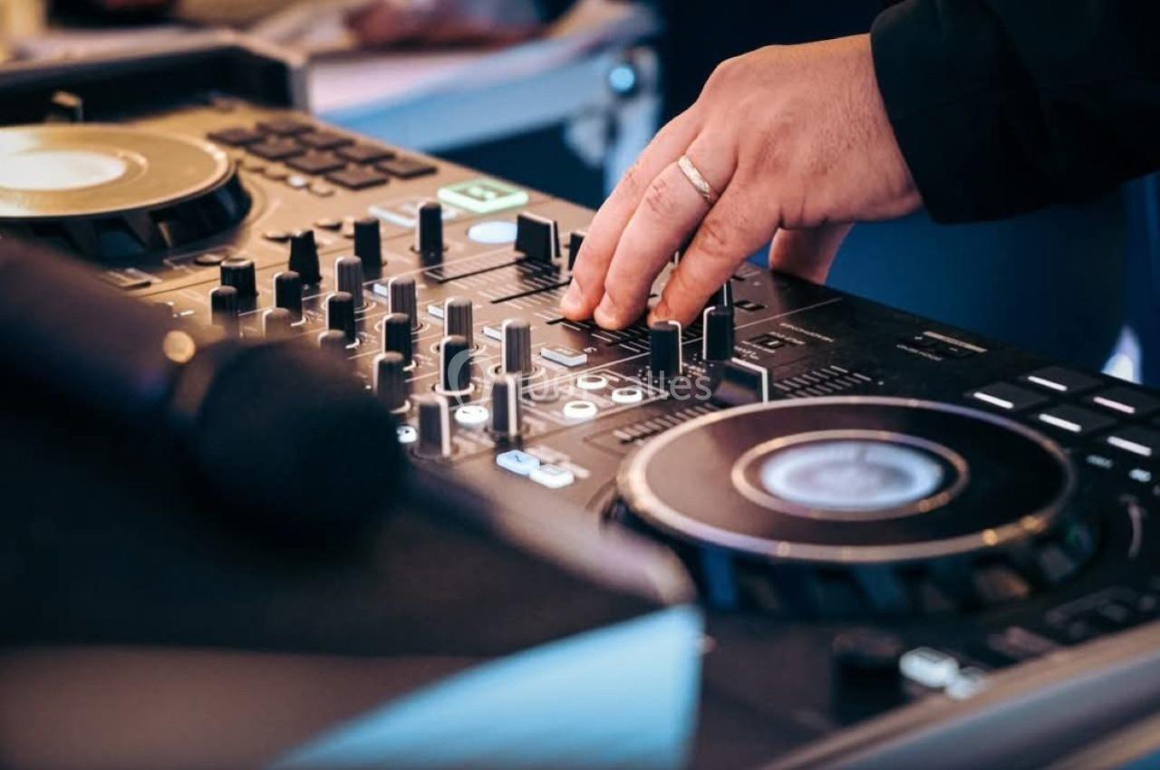 Main posée sur une table de mixage avec des boutons et platines, microphone flou au premier plan.