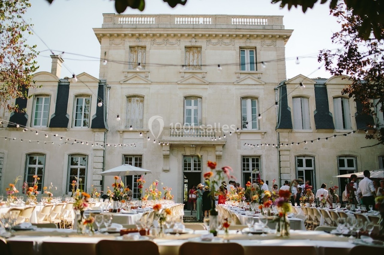 Façade d'un bâtiment élégant avec des tables décorées de fleurs pour un événement en plein air, sous des guirlandes…