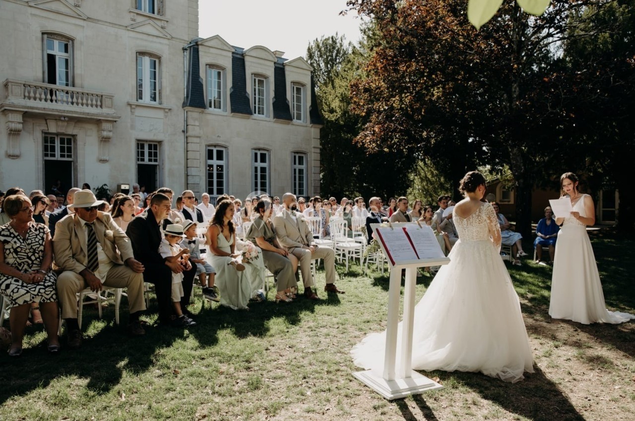Une cérémonie de mariage en extérieur devant un bâtiment ancien, avec des invités assis et deux mariées échangeant leurs…