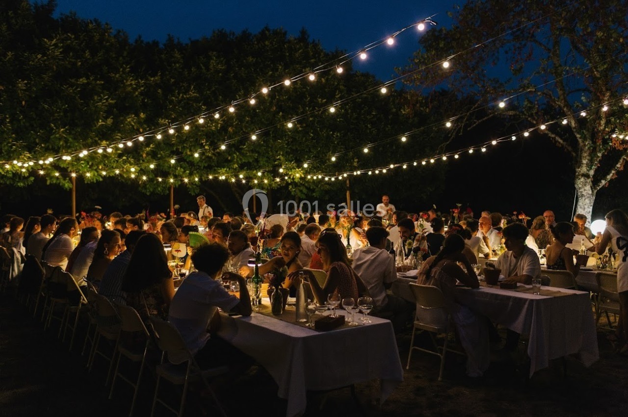 Dîner en plein air sous des guirlandes lumineuses, avec de nombreuses personnes assises à des tables longues.