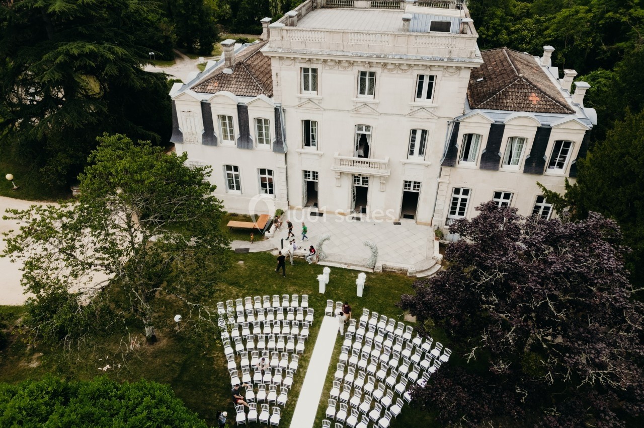 Vue aérienne d'un manoir entouré de verdure, avec une cérémonie en plein air et des chaises disposées en arc.