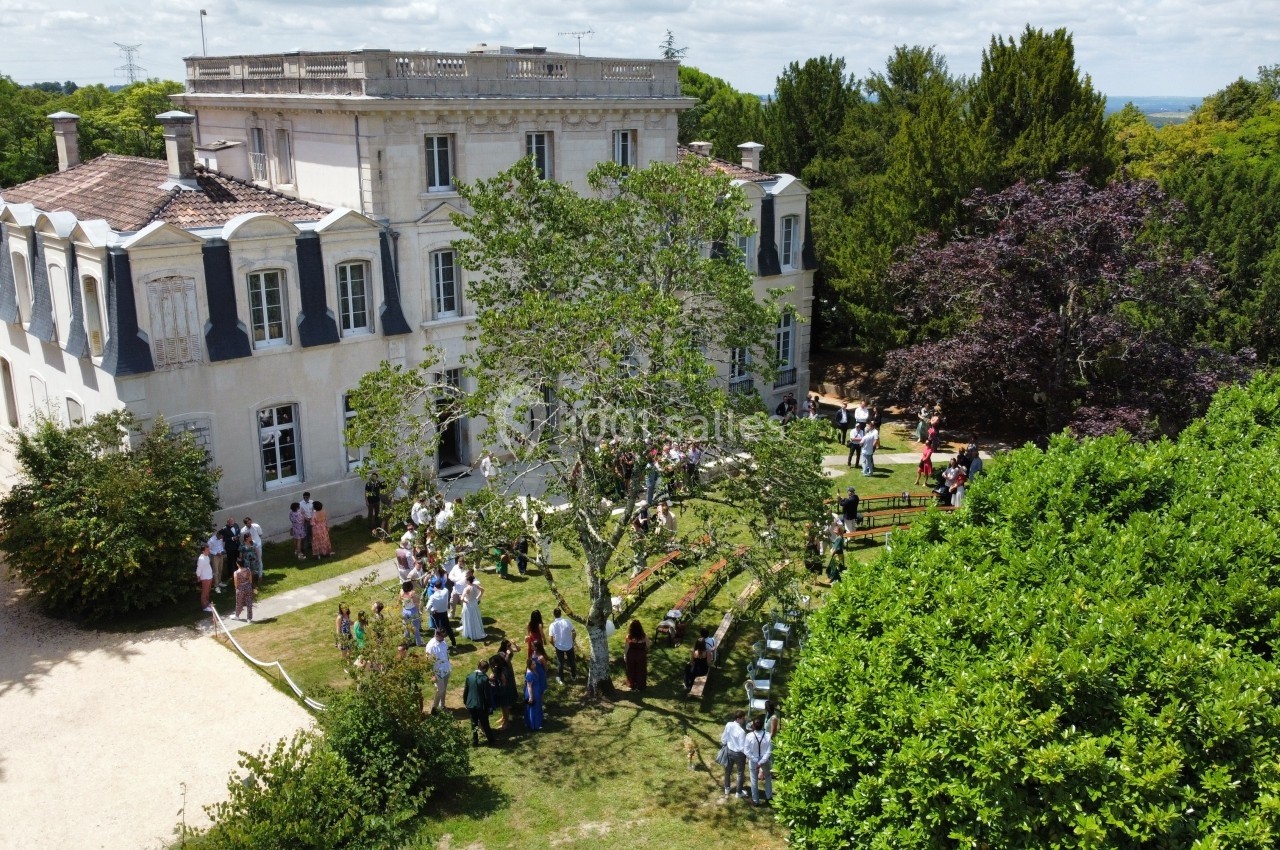 Vue aérienne d'un manoir entouré de jardins, avec des personnes rassemblées sur la pelouse lors d'un événement en plein air.