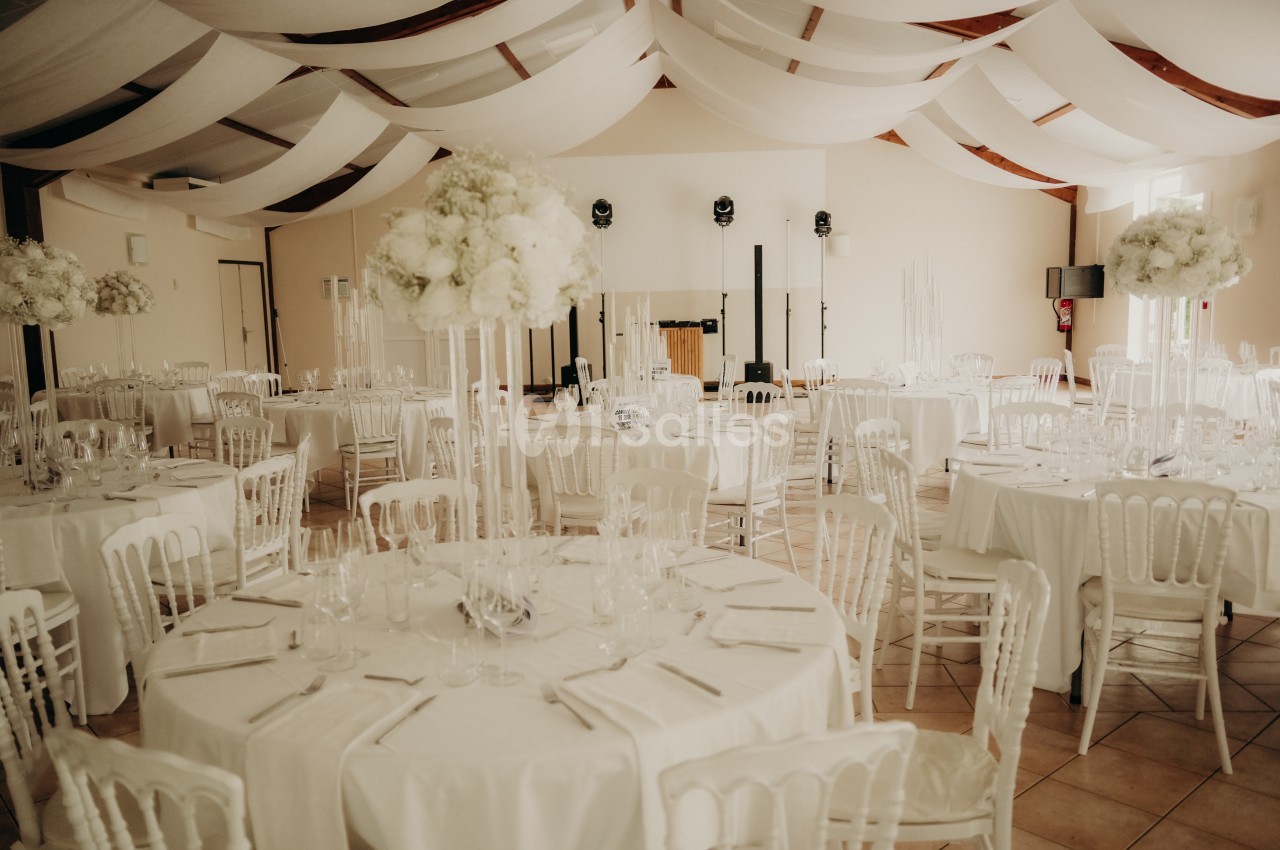 Salle de réception décorée avec des tables rondes, nappes blanches, chaises assorties et arrangements floraux élégants.