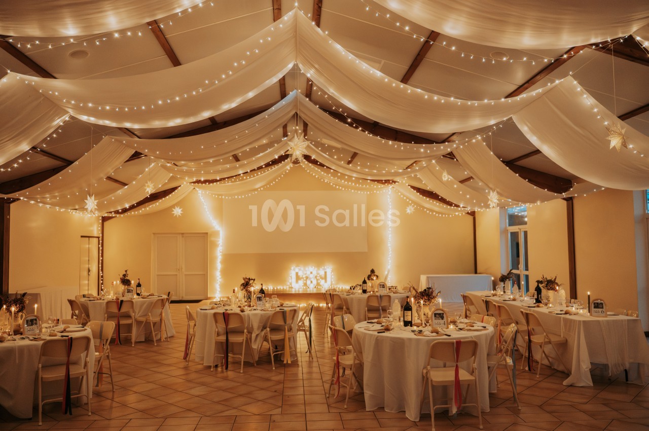 Salle de réception décorée avec des guirlandes lumineuses, des tables rondes dressées et un plafond drapé de tissu blanc.