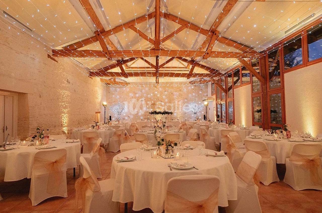 Salle de réception décorée avec des tables rondes, nappes blanches, chaises ornées et guirlandes lumineuses suspendues.