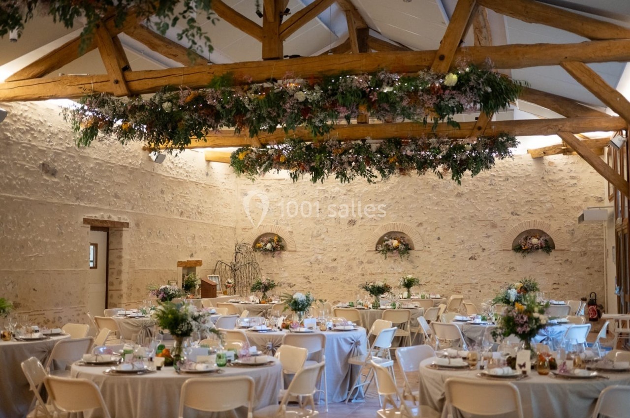 Salle de réception décorée avec des tables dressées, des fleurs suspendues et des murs en pierre apparente.