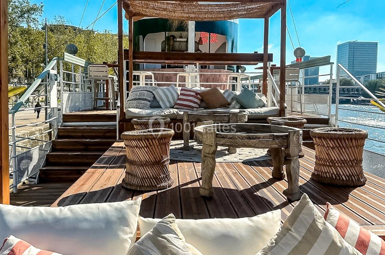 Espace extérieur d'un bateau aménagé avec des coussins, des fauteuils et une pergola en bois, sous un ciel ensoleillé.