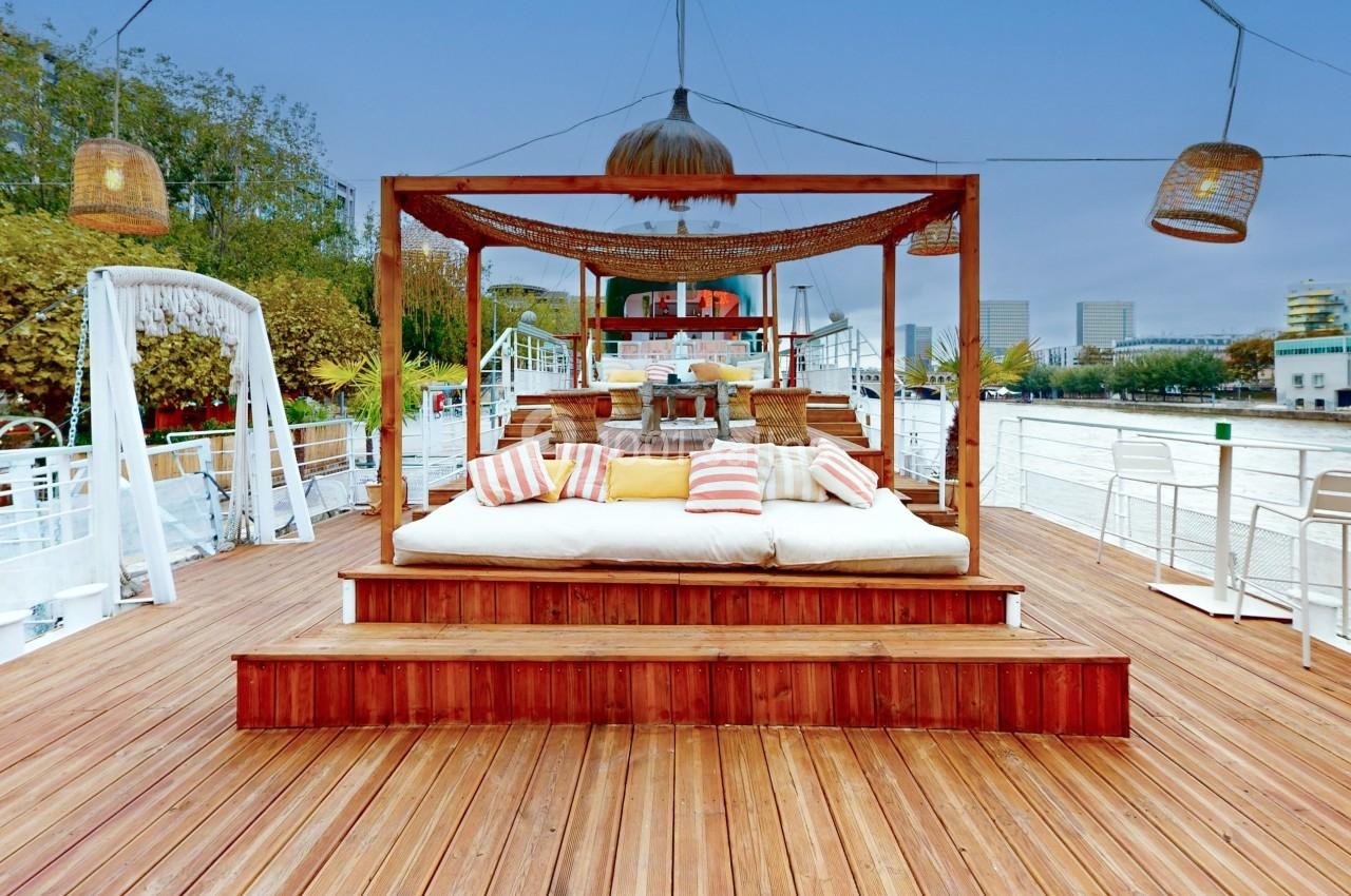 Lit extérieur avec coussins colorés sous une pergola en bois, installé sur une terrasse en bord de rivière.