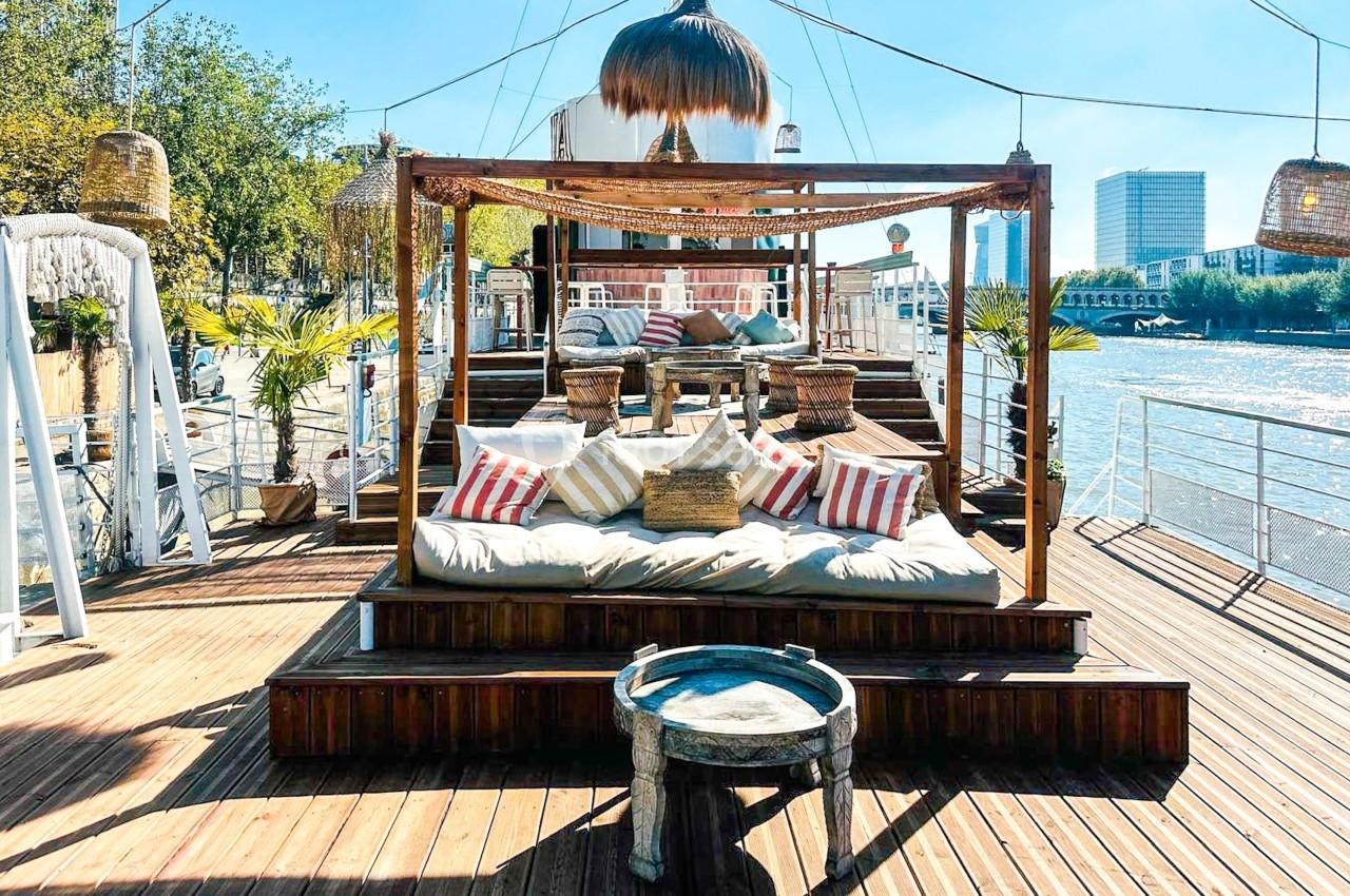 Espace détente extérieur sur une péniche avec coussins, voilages, plantes et vue sur un fleuve en arrière-plan.