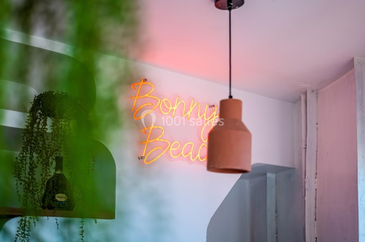 Enseigne lumineuse ’Bonny Beach’ au néon orange, suspendue dans un intérieur avec plantes et lampe en céramique.