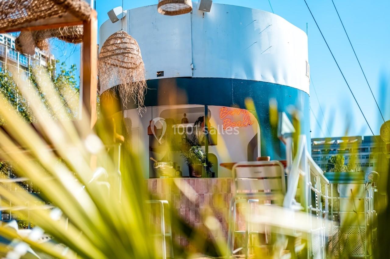 Vue d'un café extérieur avec des plantes et des lampes suspendues, partiellement masqué par des feuilles au premier plan.