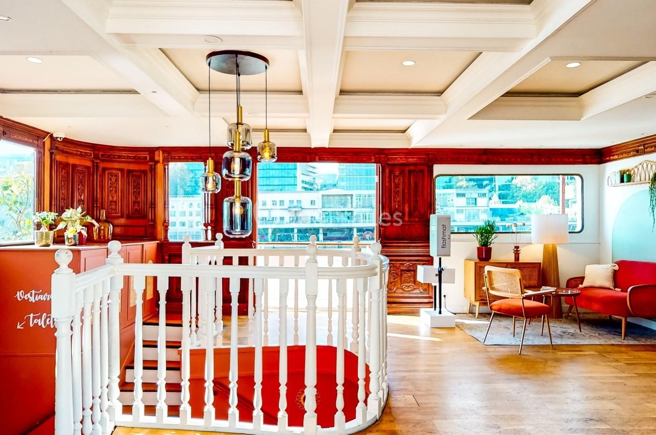 Intérieur lumineux avec escalier blanc, boiseries, fauteuils rouges et grandes fenêtres donnant sur des immeubles modernes.