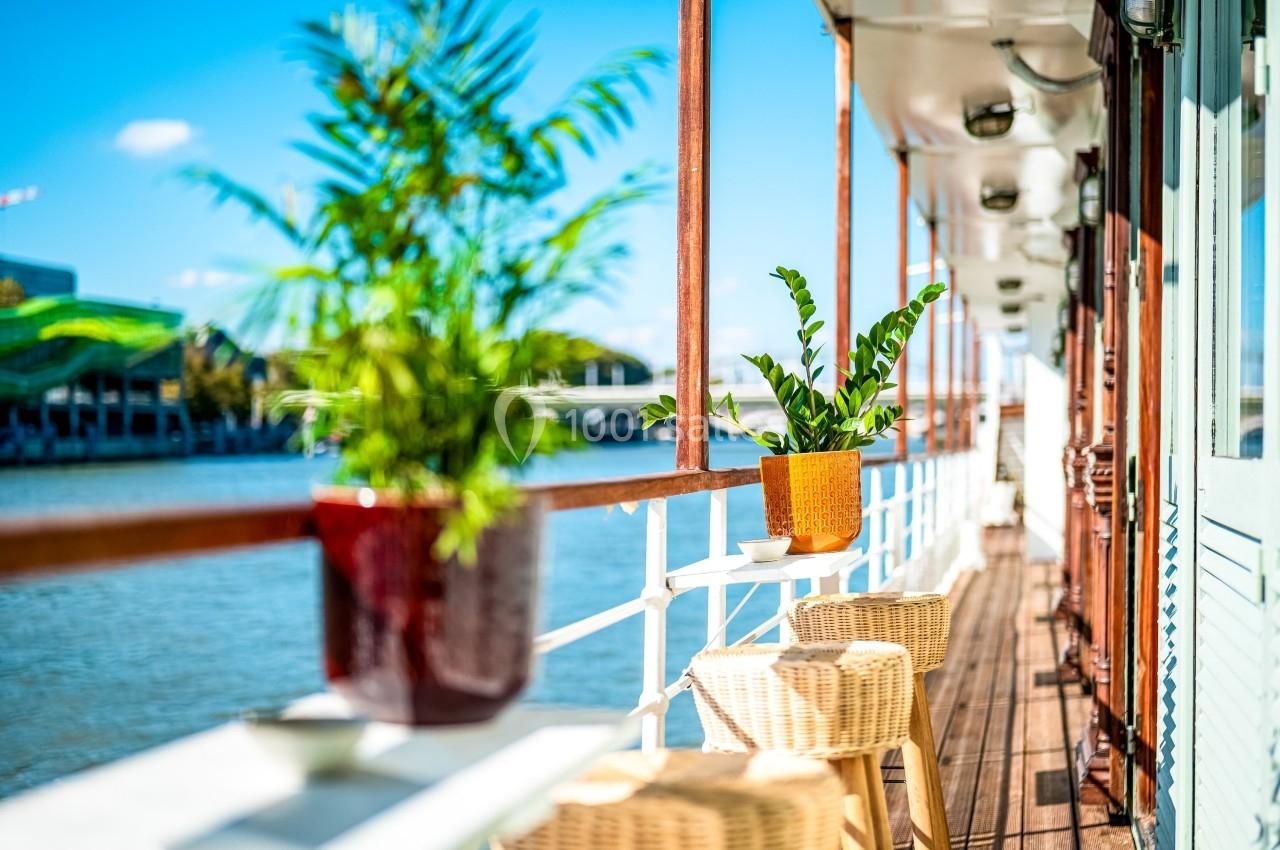 Terrasse en bois d'un bateau avec des plantes en pot, des tabourets en osier et vue sur une rivière par temps ensoleillé.