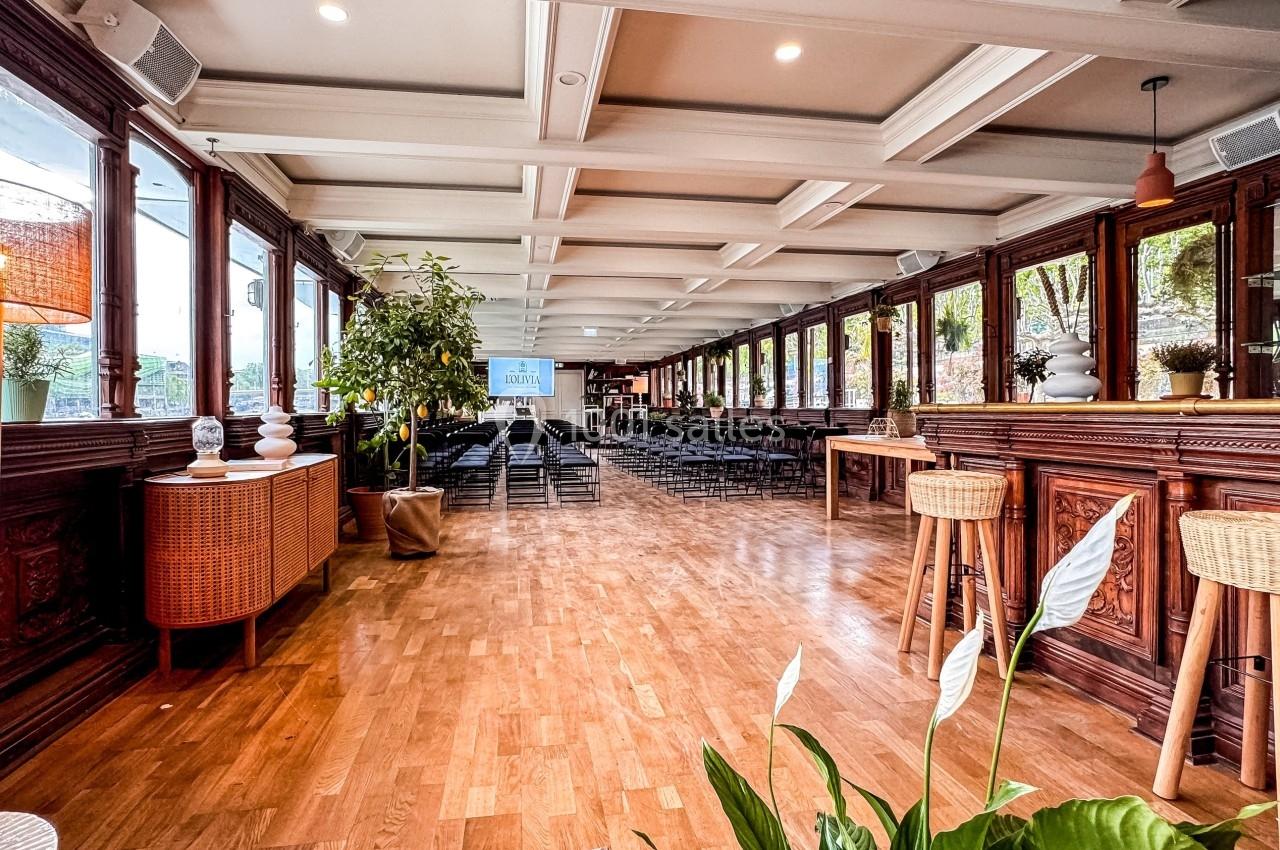 Salle lumineuse avec parquet, grandes fenêtres, plantes décoratives et chaises disposées pour un événement.