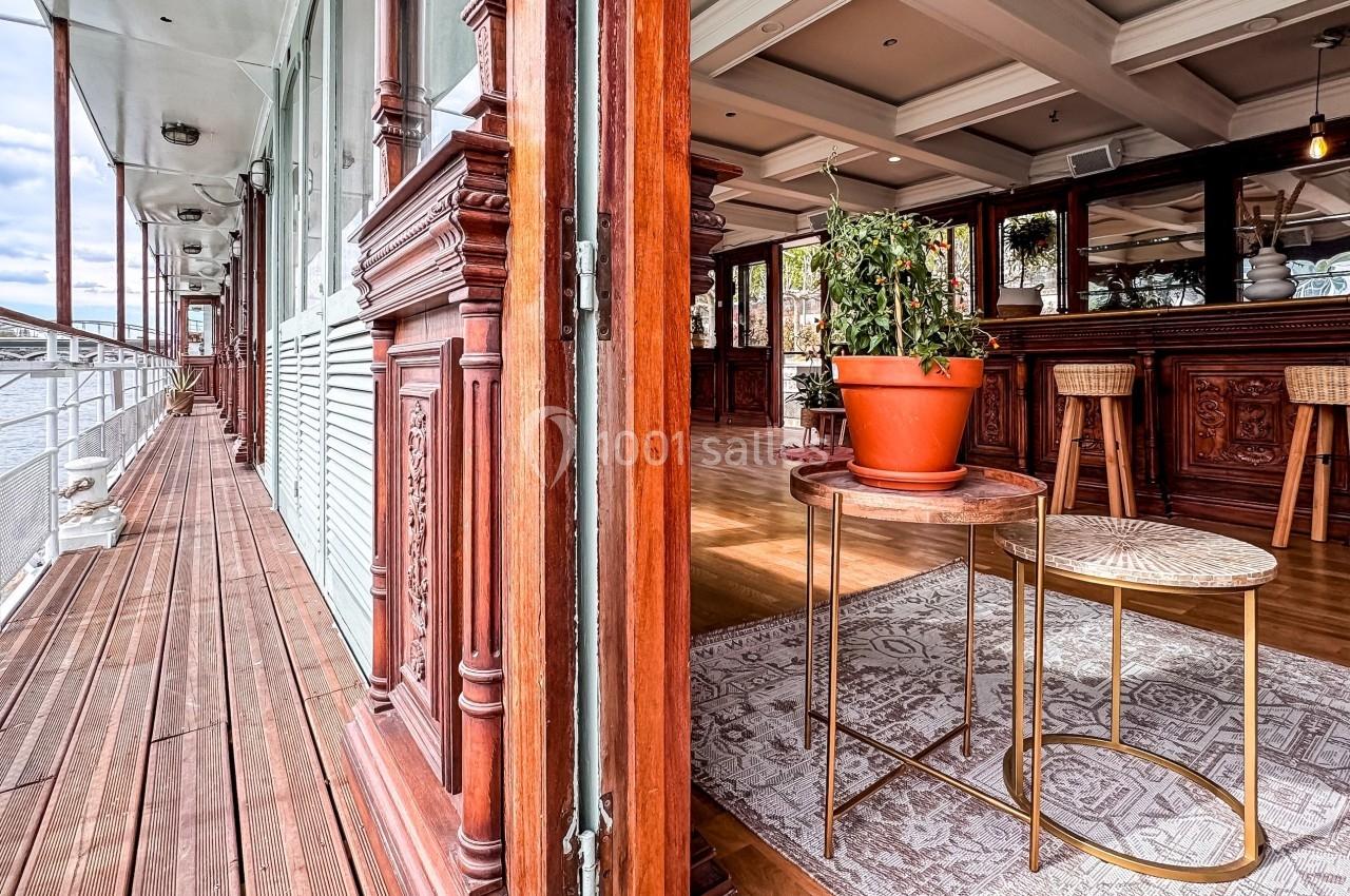 Intérieur lumineux d'un bateau avec mobilier en bois sculpté, plantes en pot et vue sur une terrasse extérieure en bois.