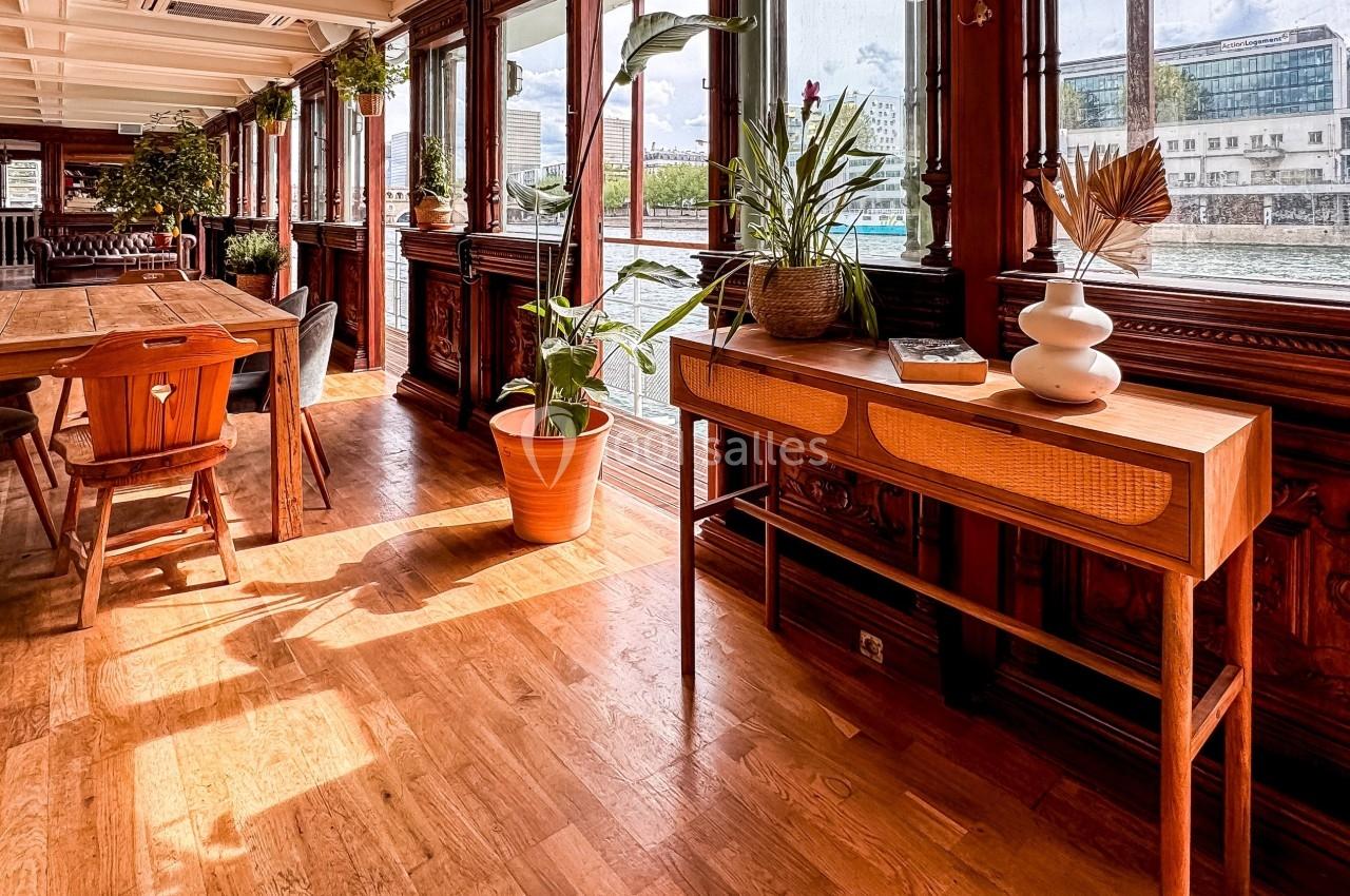 Salle lumineuse avec parquet, meubles en bois, plantes et vue sur un cours d'eau à travers de grandes fenêtres.