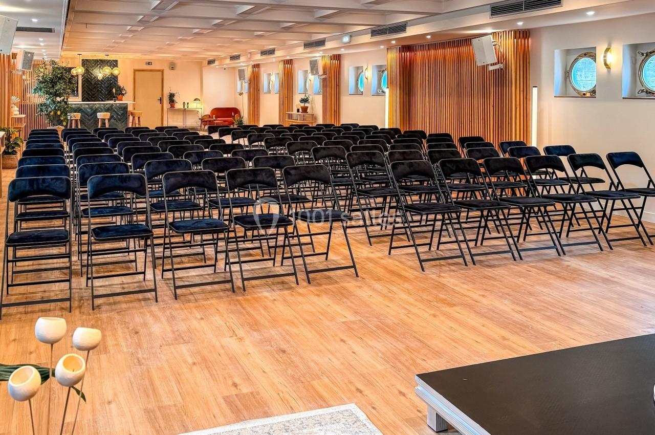 Salle lumineuse avec des rangées de chaises noires alignées, parquet en bois et décorations modernes.