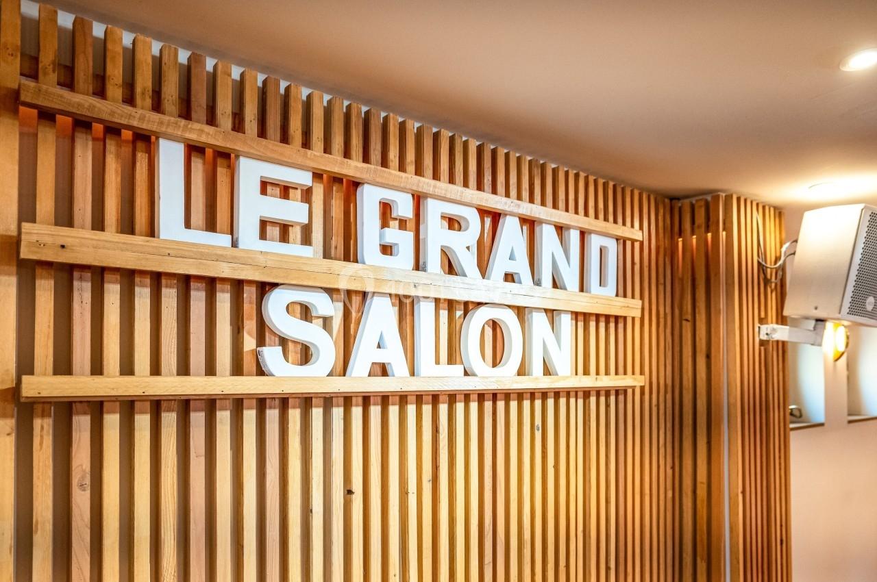 Mur en bois avec des lettres blanches formant les mots ’Le Grand Salon’, éclairé par des lumières chaudes.