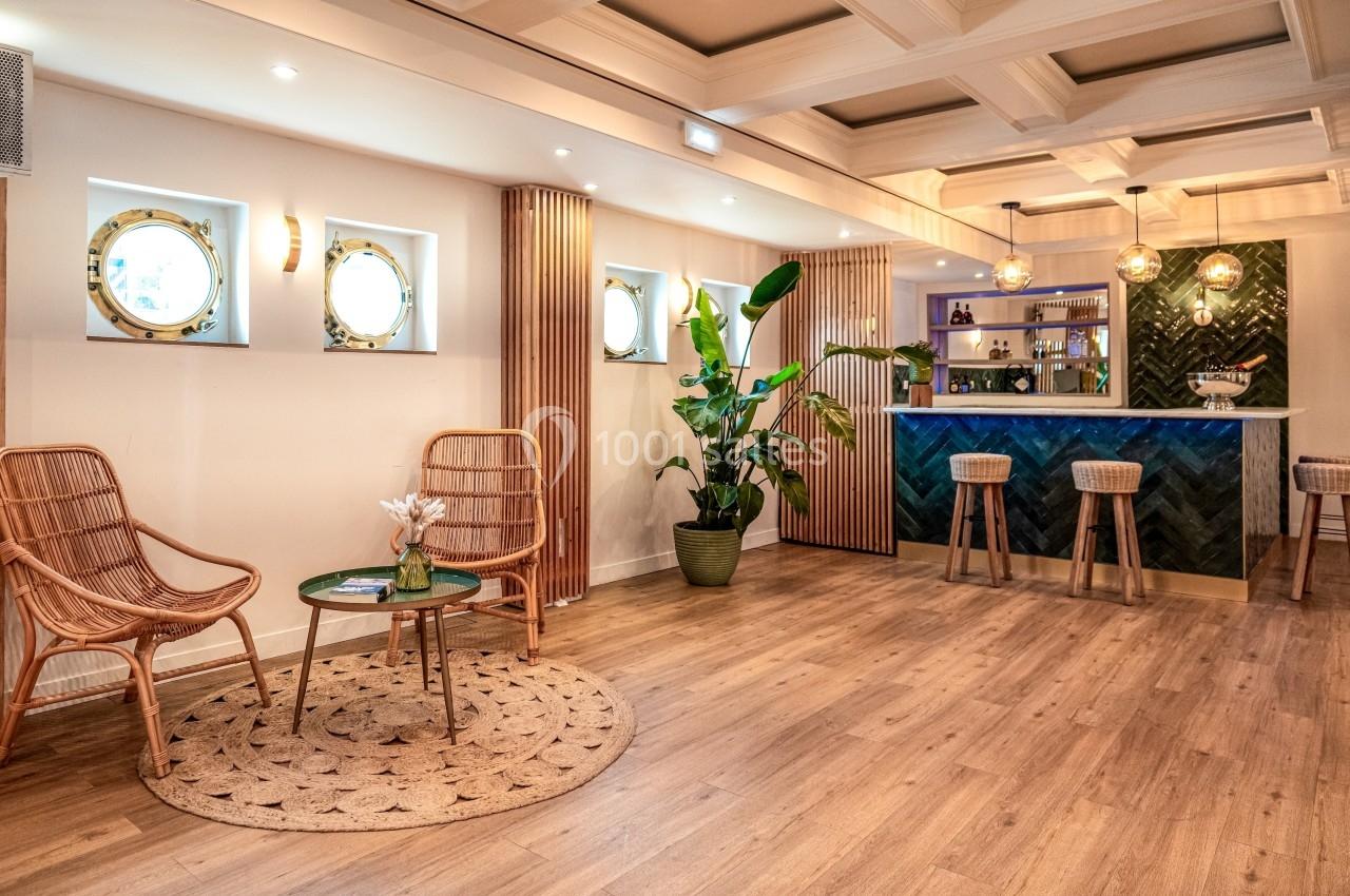 Espace intérieur lumineux avec bar en bois, tabourets, fauteuils en rotin, plantes et hublots décoratifs sur le mur.