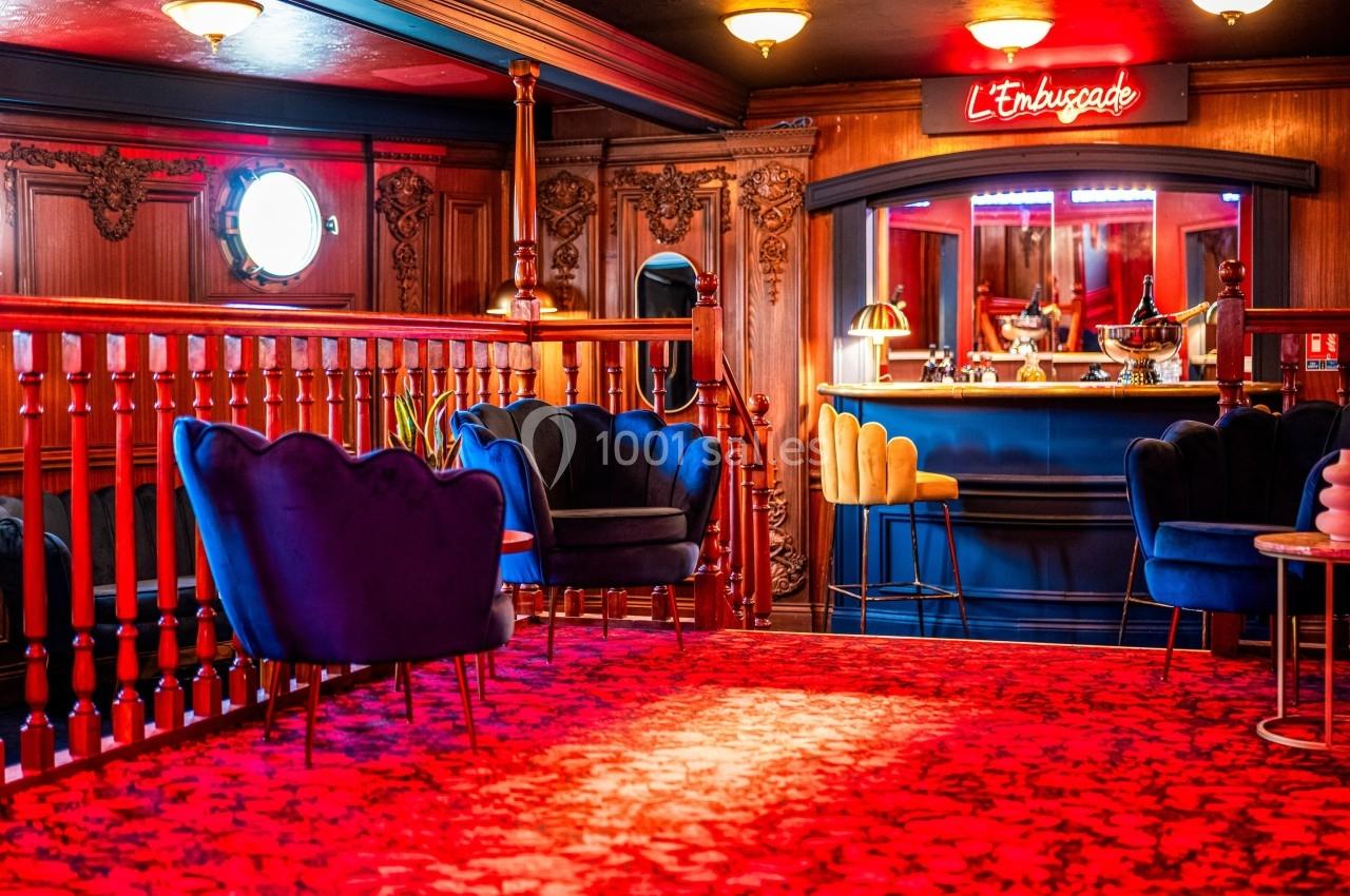 Espace lounge avec fauteuils bleus, tapis rouge à motifs et bar éclairé par une enseigne néon rouge.