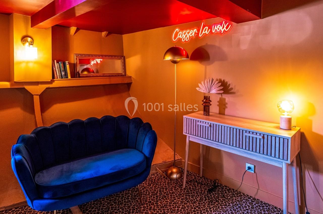 Salon cosy avec canapé bleu, lampe dorée, console blanche, miroir et néon rouge indiquant ’Casser la voix’.
