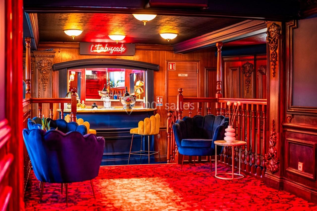 Salon chaleureux avec fauteuils bleus, bar en arrière-plan et enseigne lumineuse rouge dans un décor boisé.