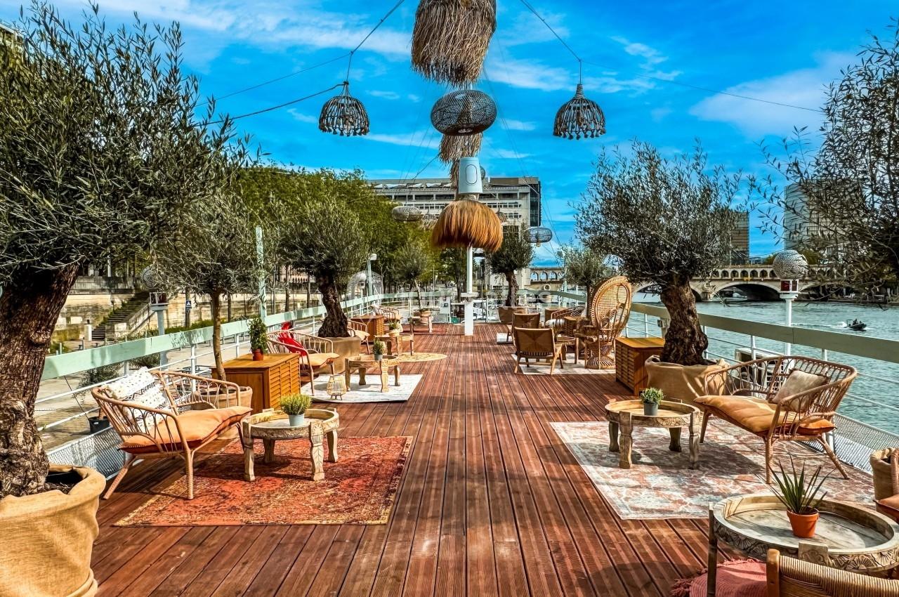 Terrasse en bois aménagée avec des fauteuils en rotin, tapis colorés, oliviers en pot et vue sur une rivière en arrière-plan.