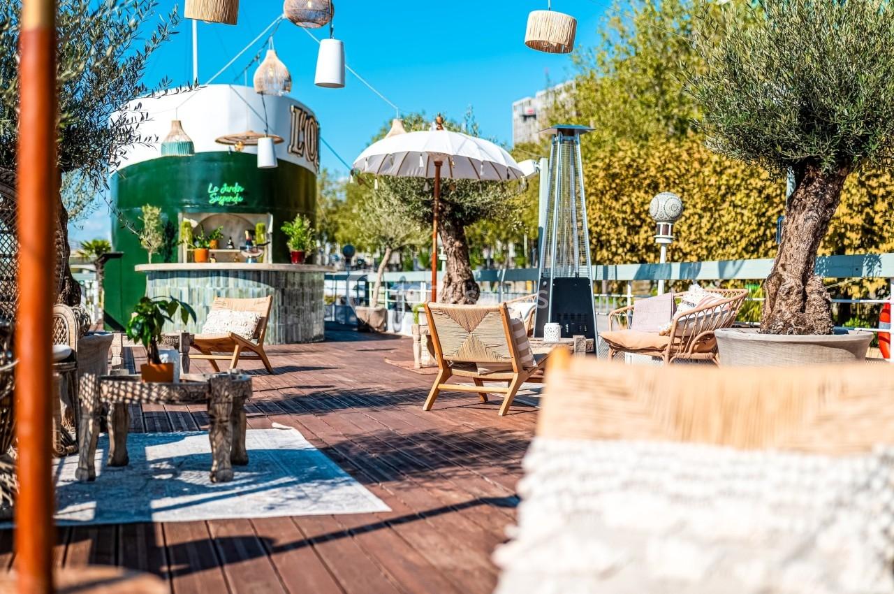 Terrasse en bois aménagée avec chaises, parasols, plantes et un comptoir de bar en arrière-plan sous un ciel bleu.