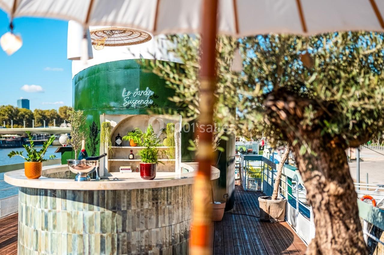 Bar extérieur en céramique verte entouré de plantes, situé sur une terrasse ensoleillée avec vue sur un fleuve.