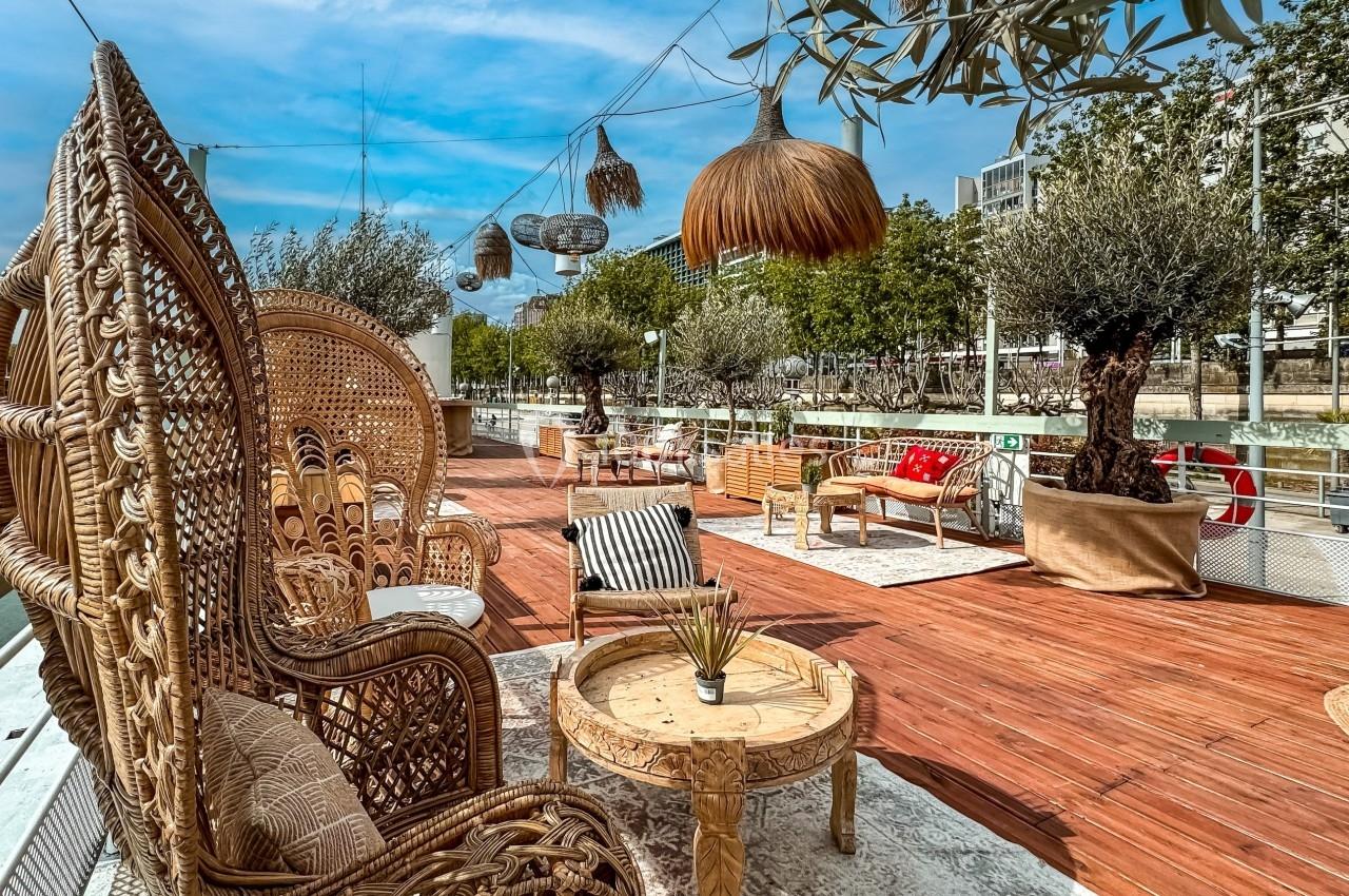 Terrasse en bois aménagée avec des fauteuils en rotin, des tables basses et des plantes, au bord d'un cours d'eau.