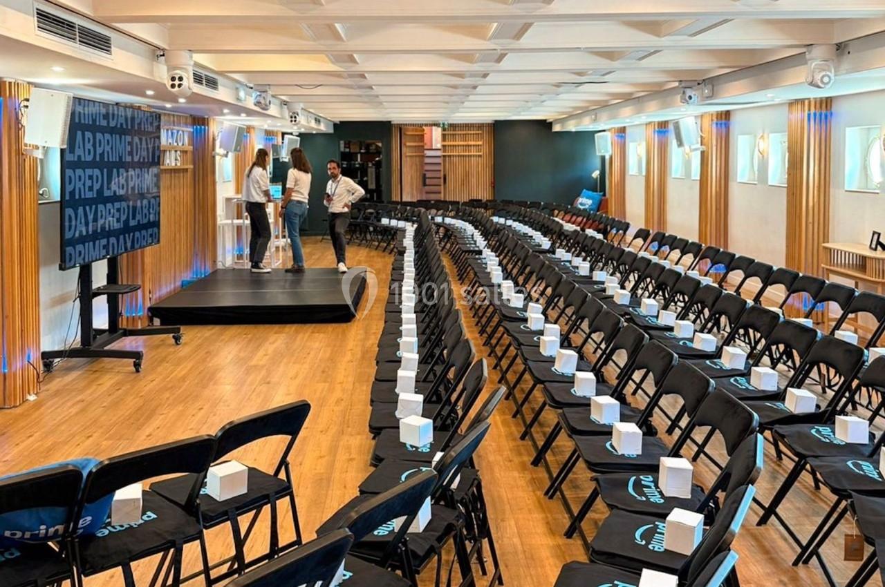 Salle de conférence avec des rangées de chaises noires alignées, chaque chaise contenant un sac blanc.