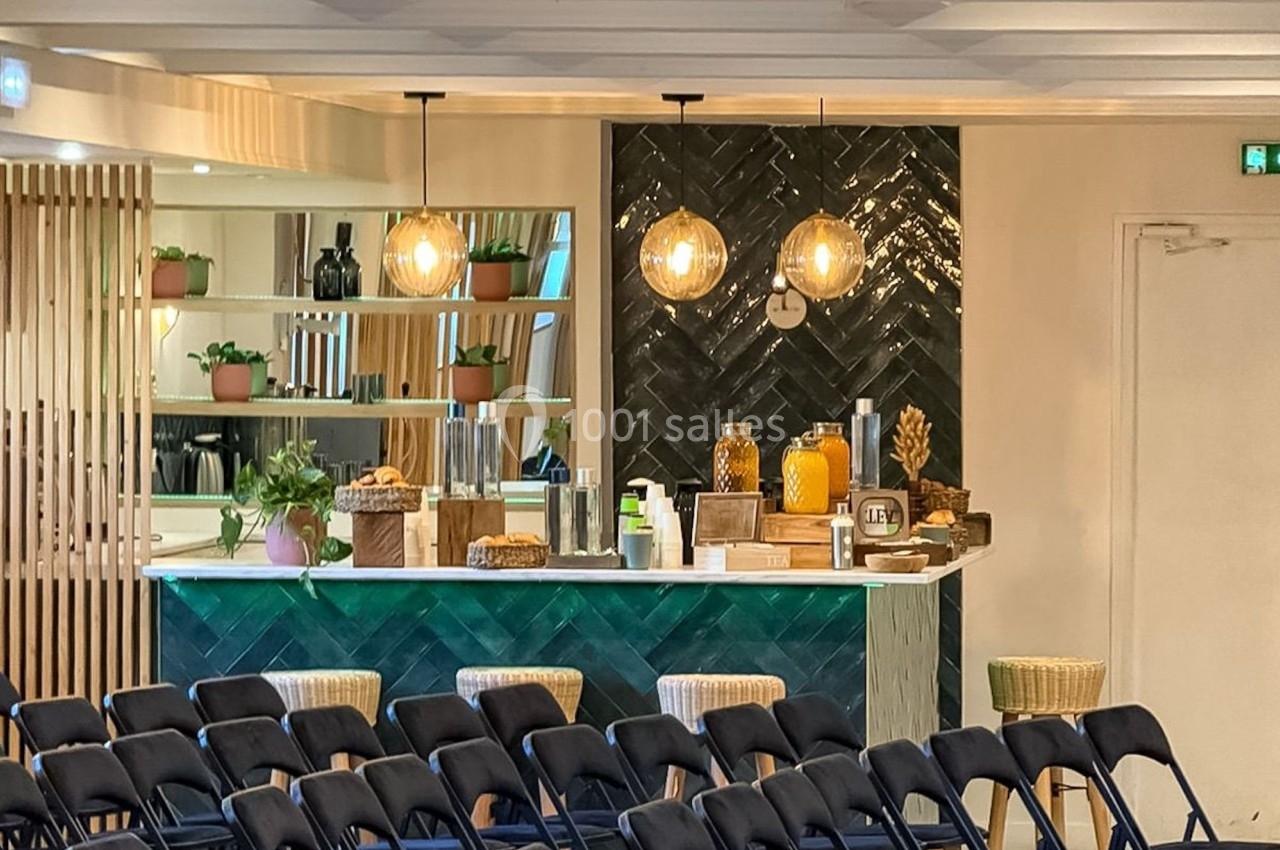 Salle aménagée avec des rangées de chaises pliantes devant un comptoir de bar décoré et éclairé par des suspensions.