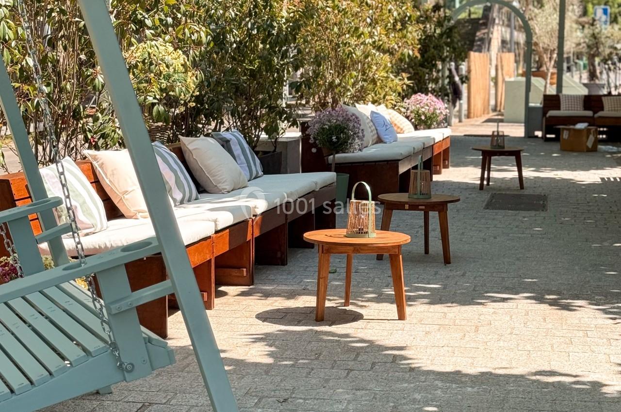 Terrasse extérieure avec bancs en bois, coussins, table basse et végétation sous un ciel ensoleillé.