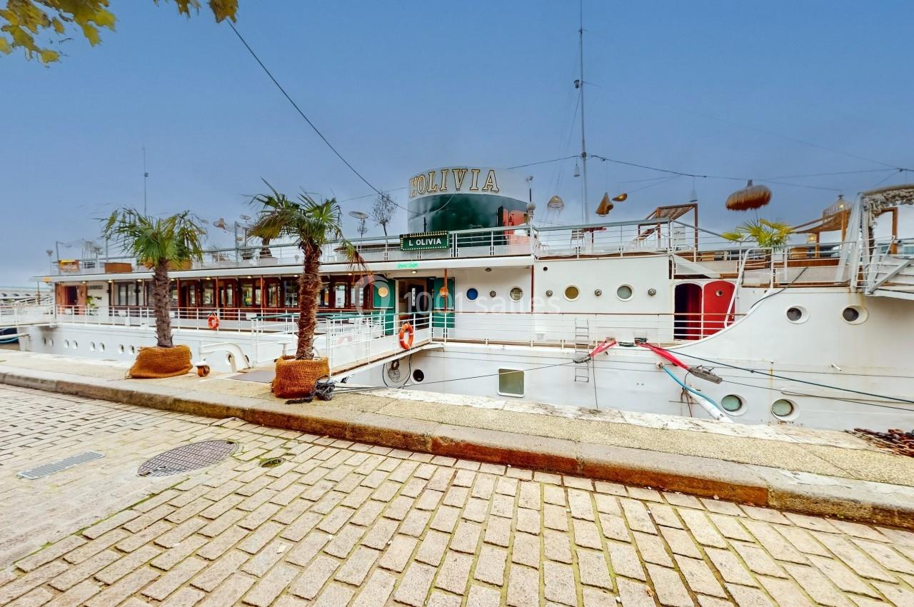 Bateau blanc amarré le long d'un quai pavé, avec des palmiers en pots et un ciel dégagé en arrière-plan.