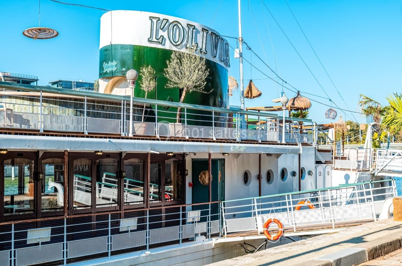 Bateau amarré avec terrasse, décorations et enseigne ’L'Olivia’, sous un ciel bleu clair.