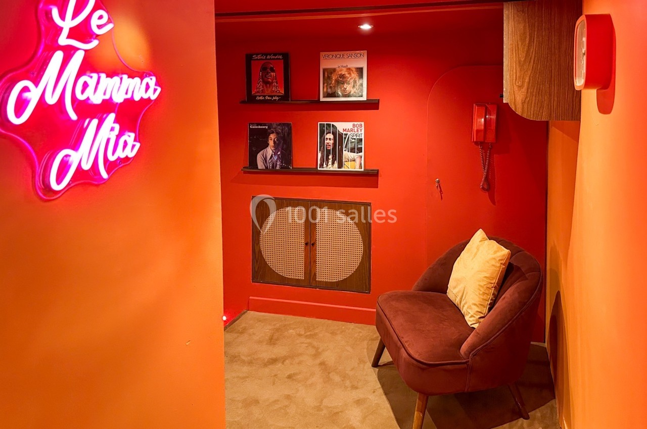 Coin cosy avec fauteuil, mur rouge orné de vinyles, téléphone vintage et enseigne néon ’Le Mamma Mia’.